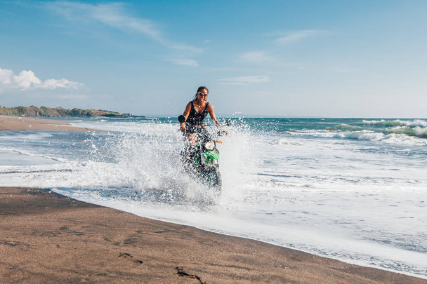 Fiatal afro-amerikai nő, lovaglás motorkerékpár enduro, fröccsenő víz beach tengerpartján  - Fotó, kép