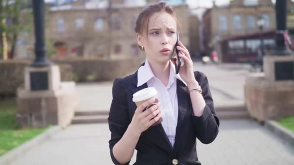 Jonge succesvolle zakenvrouw met behulp van slimme telefoon in het stadscentrum van de, professionele vrouwelijke werkgever in gesprek met zakenpartner koffie drinken. Zakelijke district wolkenkrabbers in achtergrond - Video