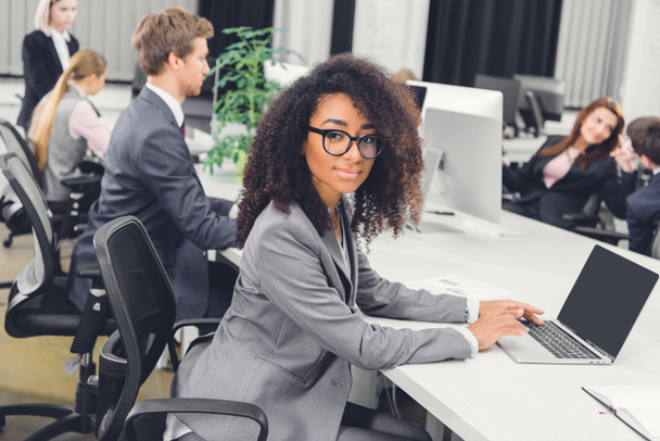 hermosa joven afroamericana mujer de negocios en gafas sonriendo a la cámara mientras se utiliza el ordenador portátil en el lugar de trabajo
  - Foto, Imagen