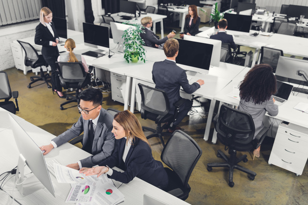 vista de ángulo alto de los empresarios multiétnicos que trabajan con papeles y computadoras de escritorio en la oficina
  - Foto, imagen