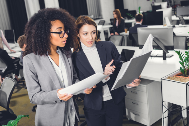 professional young multiethnic businesswomen looking at folder with papers while standing in open space office - Foto, Imagem