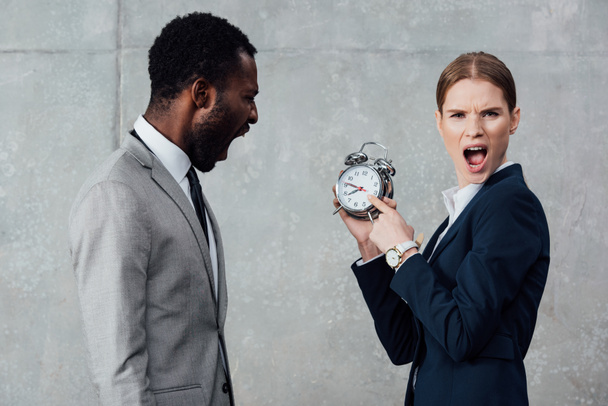donna d'affari guardando la fotocamera e indicando orologio mentre l'uomo d'affari africano americano grida su sfondo grigio
 - Foto, immagini
