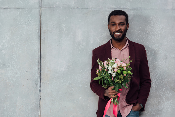 uśmiechający się african american man w ubrania dorywczo gospodarstwa kwiaty i patrząc na kamery na szarym tle - Zdjęcie, obraz