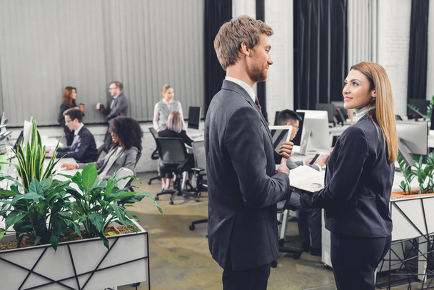 side view of young businessman and businesswoman looking at each other while standing together in open space office - Photo, Image