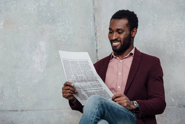 笑みを浮かべて座っているカジュアルのアフリカ系アメリカ人のビジネスマンと灰色の背景に新聞を読む - 写真・画像