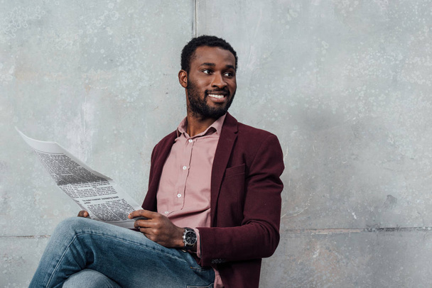 schöner lächelnder afrikanisch-amerikanischer Geschäftsmann, der auf grauem Hintergrund sitzt und Zeitung liest - Foto, Bild