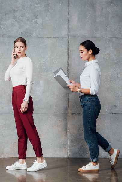 mujeres de negocios casuales multiétnicas enfocadas hablando en un teléfono inteligente y sosteniendo la carpeta con documentos en la oficina
 - Foto, Imagen
