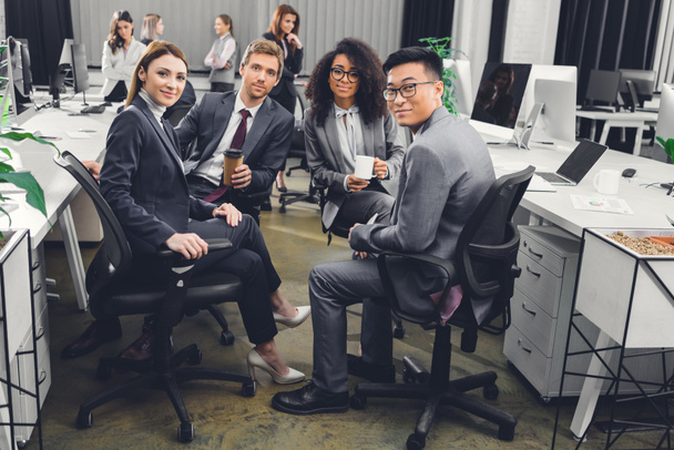 jóvenes empresarios multiétnicos en ropa formal sentados juntos y sonriendo a la cámara en la oficina
    - Foto, imagen