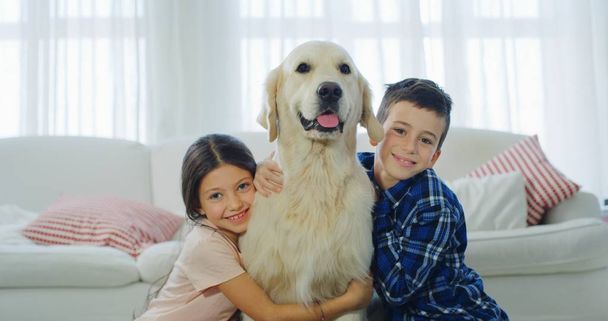 Portrait of little brother and sister cuddling their dog in living room. Concept of love for animals, childhood, pedigreed dog - Valokuva, kuva