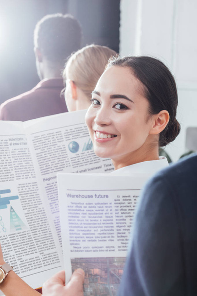 smiling asian businesswoman looking at camera and holding newspaper with colleagues on background - Foto, Imagen