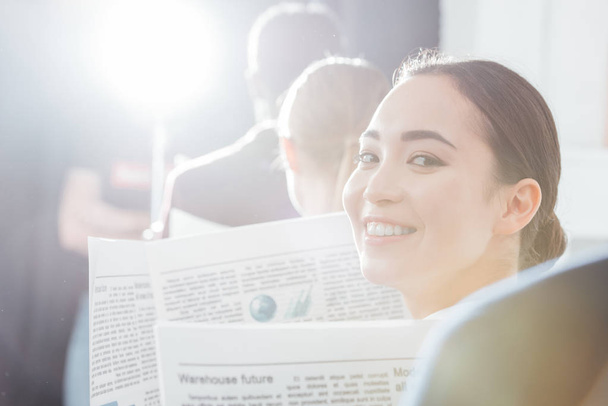 selective focus of asian businesswoman looking at camera and holding newspaper with colleagues on background - Photo, Image