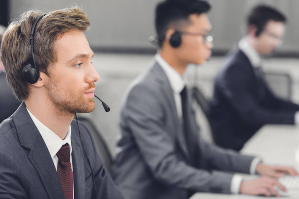 side view of focused young businessman in headset working with colleagues in call center - Φωτογραφία, εικόνα