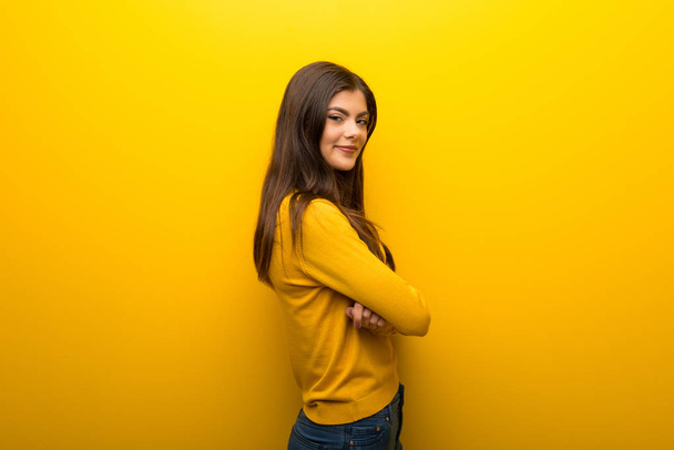 Teenager girl on vibrant yellow background keeping the arms crossed in lateral position while smiling - Photo, Image