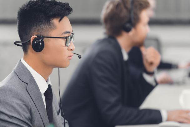 vista lateral de focado jovem asiático empresário no fone de ouvido trabalhando com colegas no escritório
 - Foto, Imagem