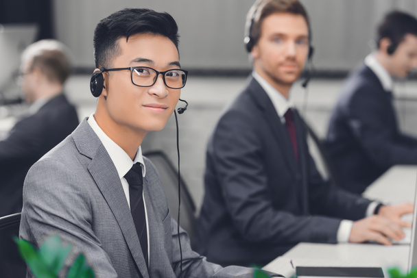 guapo joven asiático hombre de negocios en auriculares sonriendo a cámara mientras trabajando con colegas en call center
  - Foto, Imagen