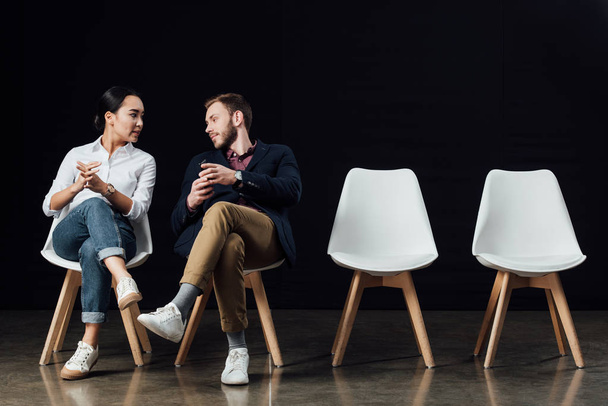 multiethnisches Paar sitzt auf Stühlen und spricht isoliert auf schwarz - Foto, Bild