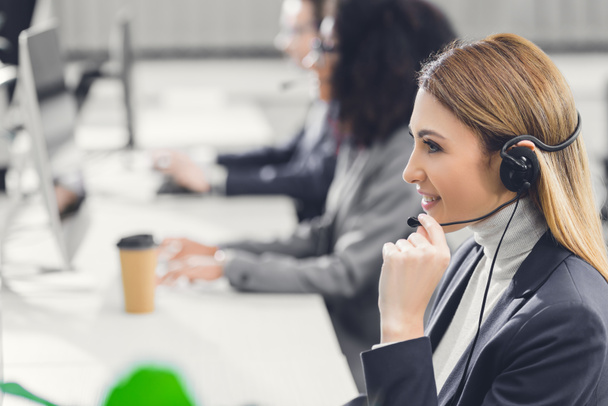 side view of smiling young businesswoman in headset working with colleagues in office  - Φωτογραφία, εικόνα