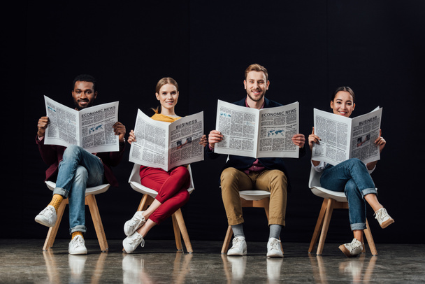 sonriente grupo multiétnico de empresarios casuales sentados en sillas y leyendo periódicos de negocios aislados en negro
 - Foto, imagen