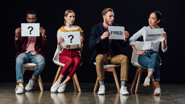 multiethnic people sitting on chairs and holding cards with 'director', 'fired' words and question marks isolated on black - Fotografie, Obrázek