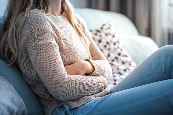 Young woman suffering from abdominal pain while sitting on sofa at home - Fotografie, Obrázek