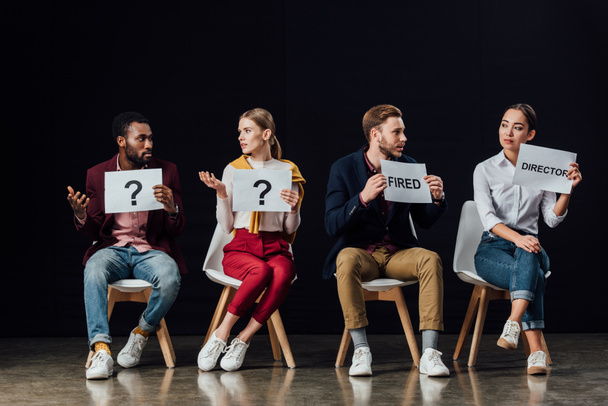 multiethnic group of people sitting and holding cards with 'director', 'fired' words and question marks isolated on black - Fotografie, Obrázek