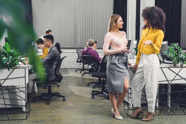 jonge multi-etnische vrouwelijke ondernemers praten en glimlachen elkaar terwijl collega's werken achter in kantoor - Foto, afbeelding