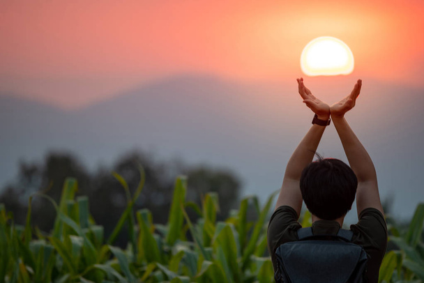 Homme asiatique dans le champ de maïs levant les bras tenant le soleil au coucher du soleil. Espérons et prions. Concept de réalisation
 - Photo, image