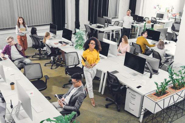 hochauflösende Ansicht von multirassischen jungen Geschäftsleuten, die im Großraumbüro mit Computern und Dokumenten arbeiten  - Foto, Bild