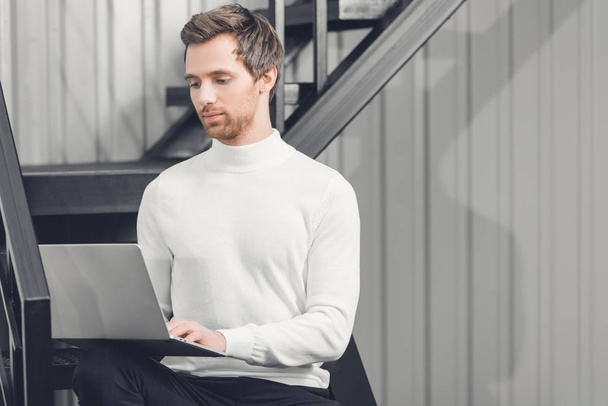 зосереджений молодий бізнесмен сидить на сходах і використовує ноутбук в офісі
  - Фото, зображення