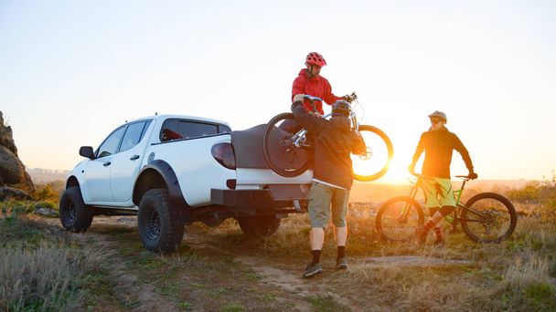 Friends Cyclists Getting Ready for Bike Riding and Taking the Bicycles off the Pickup Offroad Truck in the Mountains at Warm Autumn Sunset. MTB Adventure and Car Travel Concept. - Photo, Image