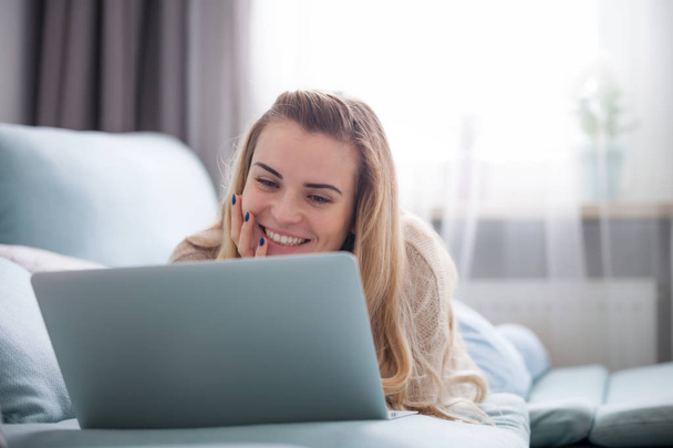 Smiling woman looking at laptop screen while lying on sofa at home, relaxing and resting - Foto, Bild