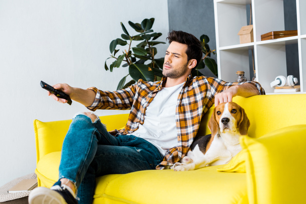 casual man with remote control watching tv and sitting on sofa with dog  - Photo, Image