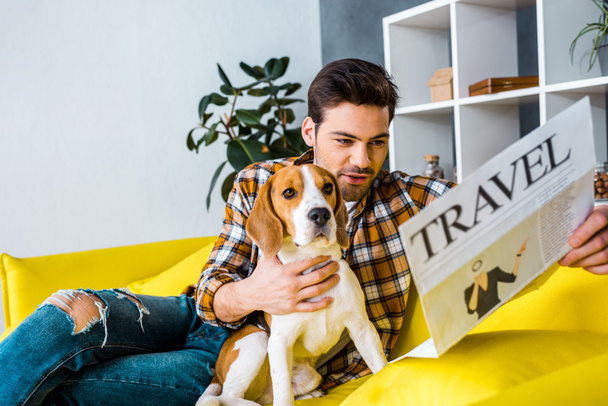 handsome man reading travel newspaper while sitting on sofa with dog - Photo, Image