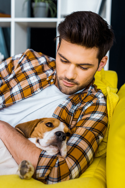 όμορφος άνδρας στο καρώ πουκάμισο ακουμπά στον καναπέ με σκύλο  - Φωτογραφία, εικόνα