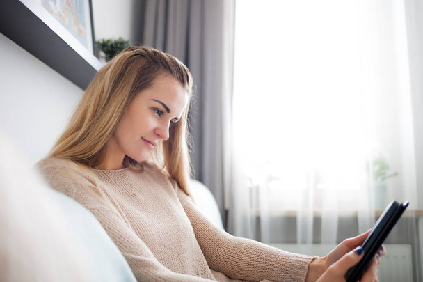 Happy woman reading ebook on digital reader while lying on comfortable sofa at home, leisure time - Φωτογραφία, εικόνα
