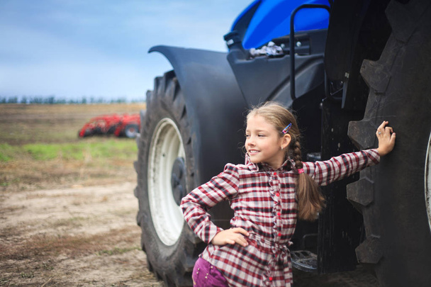 Leuk meisje in de buurt van de moderne tractor in het veld. Het begrip veldwerk, landbouwmachines. - Foto, afbeelding