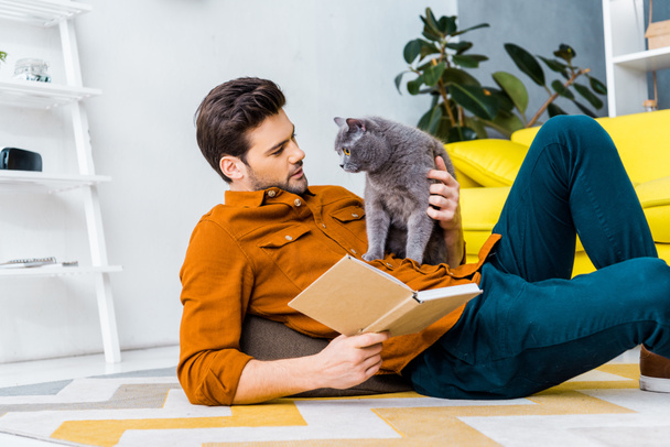 schöner Mann mit Buch und auf dem Boden liegend mit niedlicher grauer Katze  - Foto, Bild
