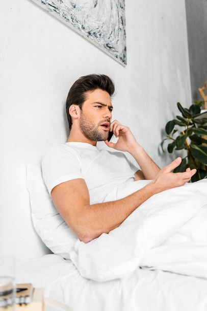 angry man talking on smartphone while resting in bed in the morning - Photo, Image