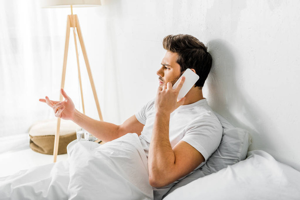 bezorgd mens gebaren en praten over smartphone zittend op bed  - Foto, afbeelding