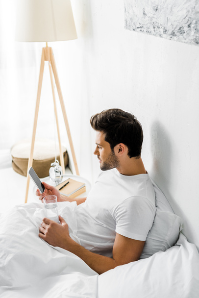 young man with glass of water using smartphone and resting in bed - Foto, Bild