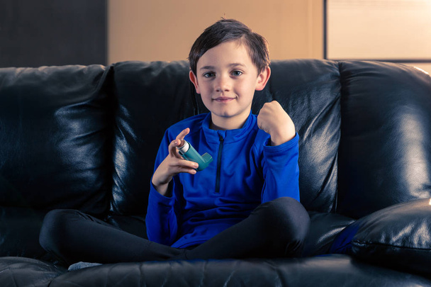 Achtjähriger Junge in sportlicher Kleidung mit Asthma-Inhalator sitzt mit geballter Faust auf Sofa. Konzepte: Gesundheit, Krankheit, Doping - Foto, Bild