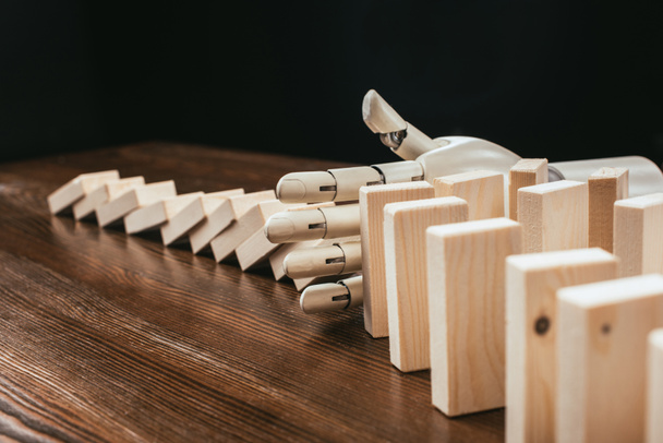 robotic hand preventing wooden blocks from falling on desk isolated on black - Foto, afbeelding