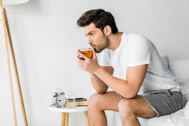 handsome man in pajamas drinking tea in bedroom - Photo, Image