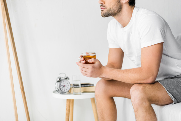 abgeschnittene Ansicht eines nachdenklichen Mannes mit einer Tasse Tee im Schlafzimmer - Foto, Bild