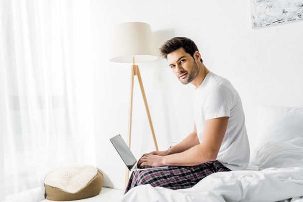 handsome man using laptop while sitting on bed in the morning - Zdjęcie, obraz