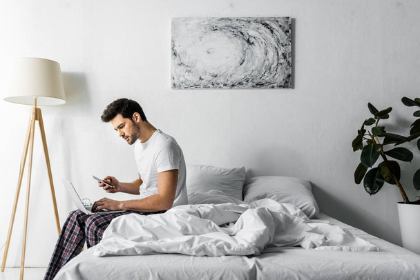 fiatal férfi pizsama használ smartphone és laptop-ban hálószoba - Fotó, kép