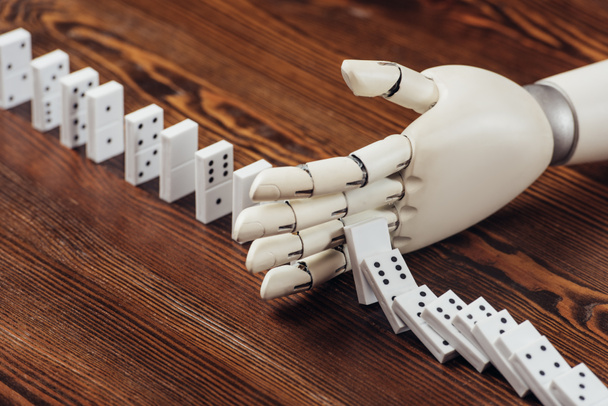 selective focus of robotic hand preventing dominoes from falling on wooden desk - Foto, Imagen