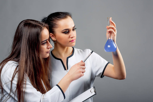 Two young pharmacists in medical clothes examining blue liquid sample on the grey background. Beautiful female women working on research. Close-up - Valokuva, kuva