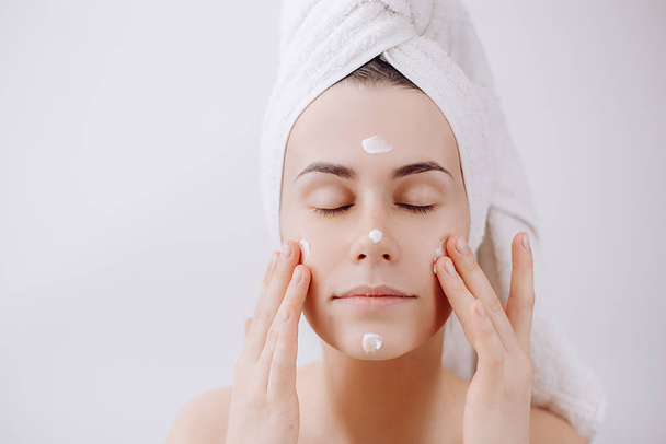 A young beautiful girl after a shower with a towel on her head apply a cleansing mask on her face. Skin care and beauty concept. Place for text - Φωτογραφία, εικόνα