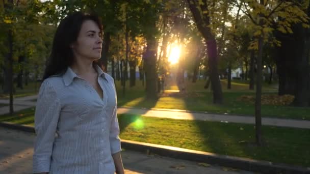 Confident Beautiful Successful Woman Walks In City Park And Thinking. Evening Sunset Background. Autumn Sunny Day In City Park. Lens Flare. Slow motion 4K 60 fps - Materiał filmowy, wideo